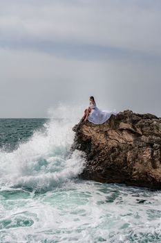 A woman in a storm sits on a stone in the sea. Dressed in a white long dress, waves crash against the rocks and white spray rises above her