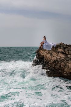 A woman in a storm sits on a stone in the sea. Dressed in a white long dress, waves crash against the rocks and white spray rises