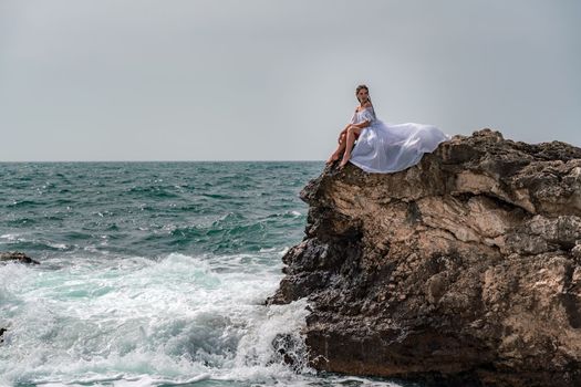 A woman in a storm sits on a stone in the sea. Dressed in a white long dress, waves crash against the rocks and white spray rises