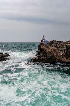 A woman in a storm sits on a stone in the sea. Dressed in a white long dress, waves crash against the rocks and white spray rises