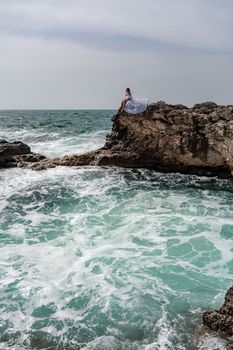 A woman in a storm sits on a stone in the sea. Dressed in a white long dress, waves crash against the rocks and white spray rises