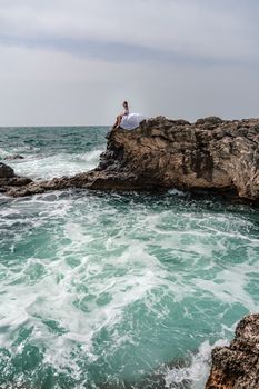 A woman in a storm sits on a stone in the sea. Dressed in a white long dress, waves crash against the rocks and white spray rises