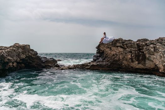 A woman in a storm sits on a stone in the sea. Dressed in a white long dress, waves crash against the rocks and white spray rises