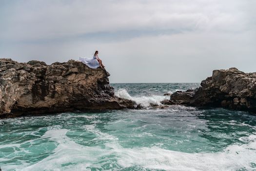 A woman in a storm sits on a stone in the sea. Dressed in a white long dress, waves crash against the rocks and white spray rises