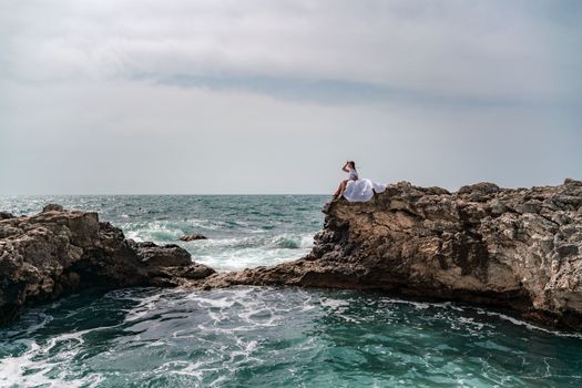 A woman in a storm sits on a stone in the sea. Dressed in a white long dress, waves crash against the rocks and white spray rises