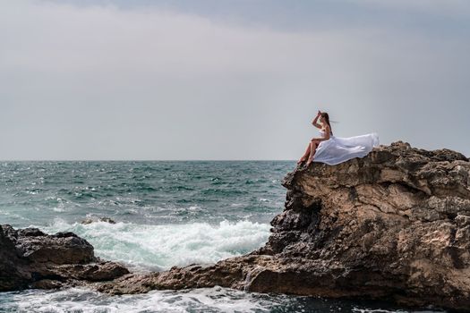 A woman in a storm sits on a stone in the sea. Dressed in a white long dress, waves crash against the rocks and white spray rises