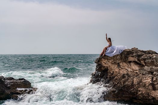 A woman in a storm sits on a stone in the sea. Dressed in a white long dress, waves crash against the rocks and white spray rises