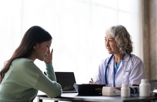 Woman senior doctor is Reading Medical History of Female Patient and Speaking with Her During Consultation in a Health Clinic. Physician in Lab Coat Sitting Behind a Laptop in Hospital Office.