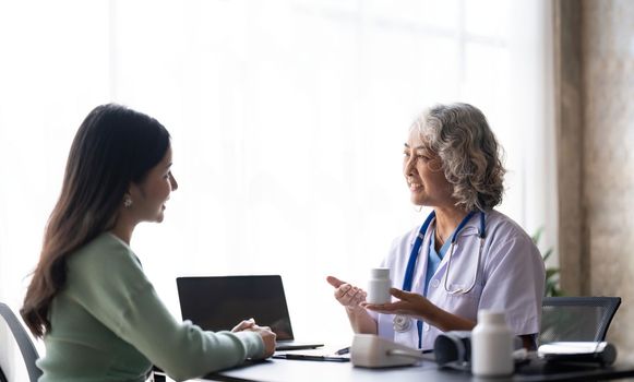 Woman senior doctor is Reading Medical History of Female Patient and Speaking with Her During Consultation in a Health Clinic. Physician in Lab Coat Sitting Behind a Laptop in Hospital Office.