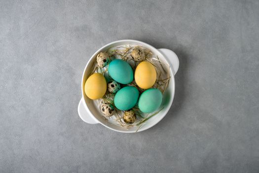 Easter composition with colored chicken eggs and quail eggs in a white dish on a gray background. View from above.