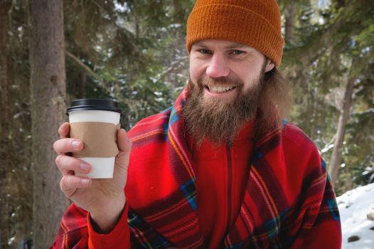Walk with a cup of hot cocoa in the winter park. Portrait of a handsome bearded guy in winter clothes in the snow drinking hot tea or coffee from a paper cup and enjoying a nice sunny snowy day.
