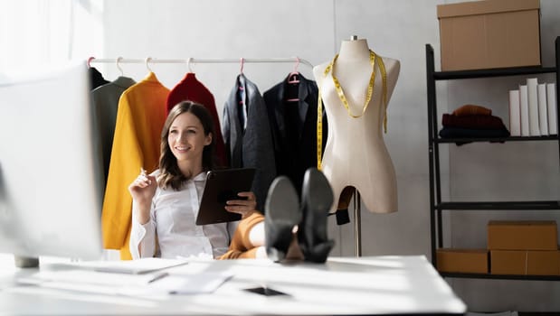 Full length portrait of attractive smiling young stylish fashion clothes designer sittng near table with computer, enjoying working in modern showroom atelier, casual workday concept...