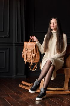 a brunette girl in a knitted beige dress poses while sitting with a leather backpack in her hands