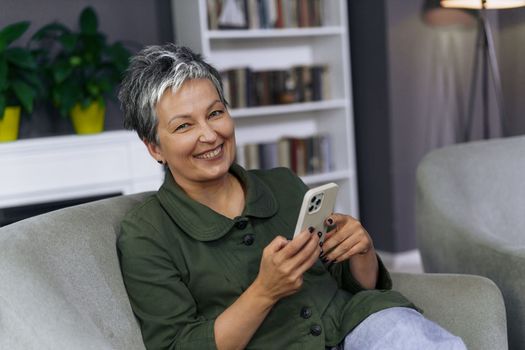 Cheerful mature woman is sitting comfortably on a sofa at home, holding a mobile phone with a bright smile on her face. She appears to be engaged in a conversation or browsing through the phone, enjoying her leisure time indoors.