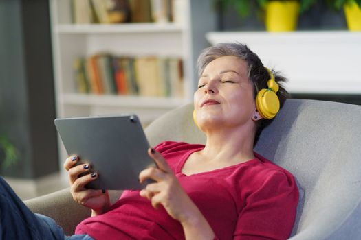 Mature woman captured on sofa home holding tablet PC and wearing headphones while listening music with her eyes closed. Woman enjoys her leisure time while indulging in pleasures of modern technology.