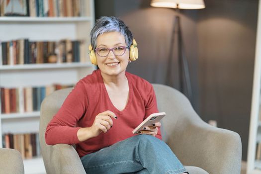 Senior woman is happily texting on her phone while wearing pink glasses and yellow headphones at home, reflecting a relaxed and modern lifestyle with a focus on technology and communication. High quality photo