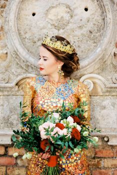 Bride in a golden crown with a bouquet in her hands stands near a wall with stucco. High quality photo