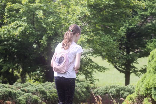 School-age girl with pink backpack walking in summer park.
