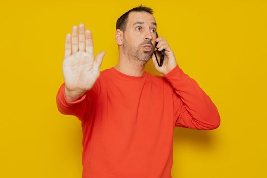 Hispanic man with beard talking on mobile phone pissed off making a stop sign with hand isolated over yellow background