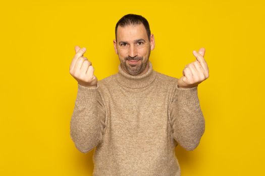 Hispanic man with a beard wearing a turtleneck making the money gesture with his fingers isolated over yellow background
