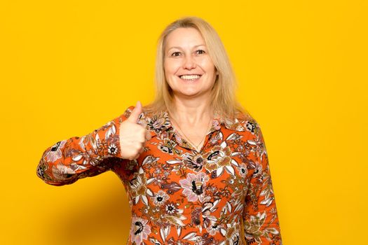 Photo portrait of nice caucasian woman with bright smile showing thumb up in patterned dress isolated on yellow colored background