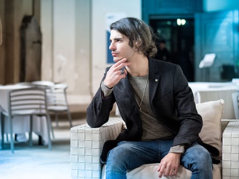 Handsome young man, wearing elegant coat posing at bar table outside on urban background.