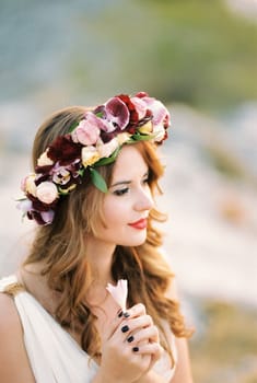 Bride in a multi-colored wreath holds a flower in her hands in front of her chest. Profile. High quality photo