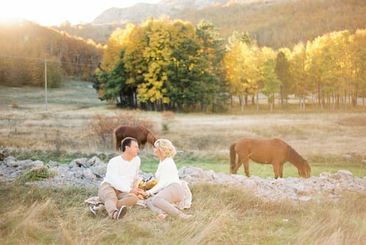 Man and woman sitting on a blanket on a lawn near grazing horses. High quality photo