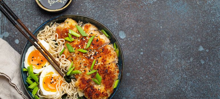 Asian noodles ramen soup with deep fried panko chicken fillet and boiled eggs in ceramic bowl with chop sticks and soy sauce on stone rustic background top view, space for text