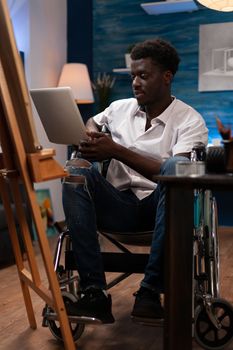 Person with artistic skills and disabilities taking online drawing courses with laptop. African american wheelchair user looking for inspiration on art web sites to create sketch masterpiece at atelier.