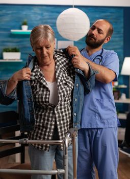 Male nurse assisting female patient in nursing home. Elderly woman using walker for walking support while specialist helps her put on jacket after physiotherapy session.