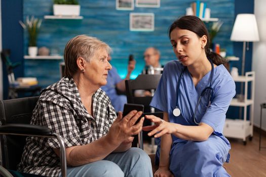 Senior female patient in wheelchair showing photos of grandchildren with smart phone to nurse. Professional health care provider attending to female resident in nursing home.