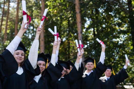 A group of graduates in robes raised their hands with diplomas outdoors. Elderly student