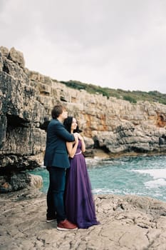 Man hugs woman from behind by the shoulders on the rocks by the sea. High quality photo