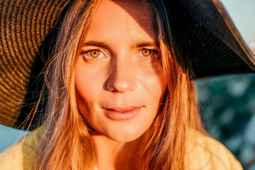 Portrait of happy young woman wearing summer black hat with large brim at beach on sunset. Closeup face of attractive girl with black straw hat. Happy young woman smiling and looking at camera at sea