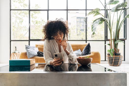 Young african american female in front of big bright window looking at cellphone eating strawberry. Home concept. Technology concept.