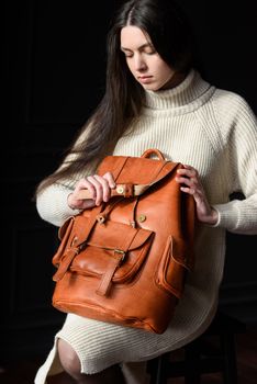 a brunette girl in a knitted beige dress poses while sitting with a leather backpack in her hands