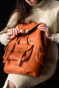 a brunette girl in a knitted beige dress poses while sitting with a leather backpack in her hands