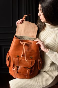 a brunette girl in a knitted beige dress poses while sitting with a leather backpack in her hands