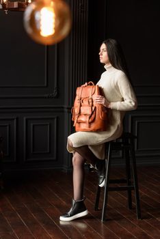 a brunette girl in a knitted beige dress poses while sitting with a leather backpack in her hands