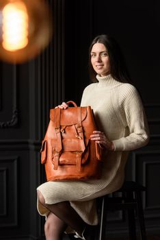 a brunette girl in a knitted beige dress poses while sitting with a leather backpack in her hands