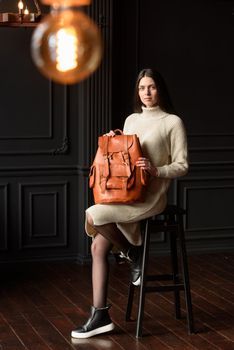 a brunette girl in a knitted beige dress poses while sitting with a leather backpack in her hands