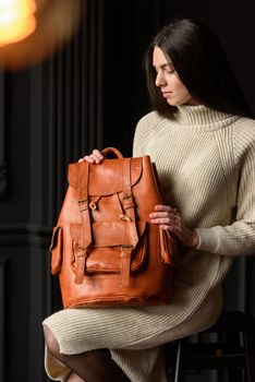 a brunette girl in a knitted beige dress poses while sitting with a leather backpack in her hands