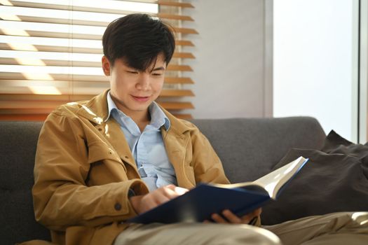 Portrait of serene asian man enjoying reading interesting book on comfortable sofa at home.