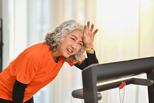Tired senior woman wiping sweat of her forehead after rinning on a treadmill indoors. Healthy active lifestyle concept.