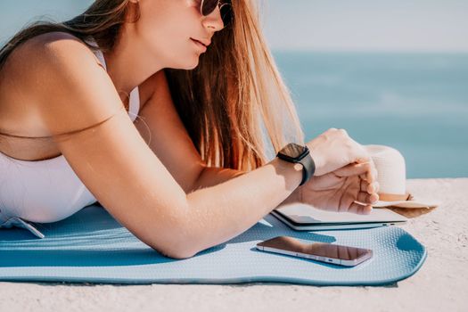 Successful business woman in yellow hat working on laptop by the sea. Pretty lady typing on computer at summer day outdoors. Freelance, travel and holidays concept.