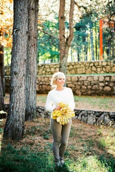 Smiling woman with a bouquet of yellow leaves stands under a tree in the park. High quality photo