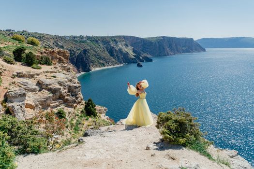 Woman in a yellow dress on the sea. Side view Young beautiful sensual woman in yellow long dress posing on a rock high above the sea at sunset. Girl in nature against the blue sky.