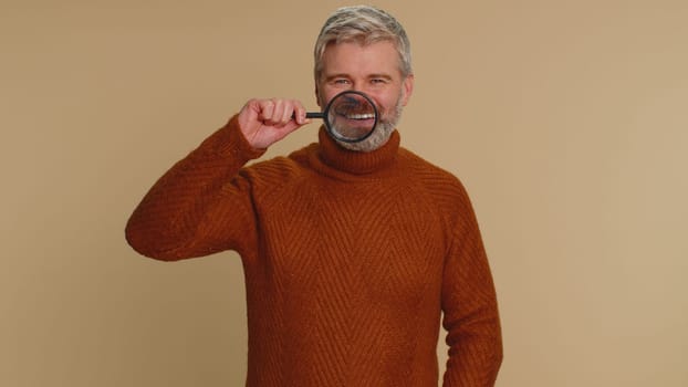 Middle-aged old man holding magnifier glass on teeth, looking at camera with happy expression, showing funny silly face smiling mouth. Senior mature adult guy isolated alone on beige studio background