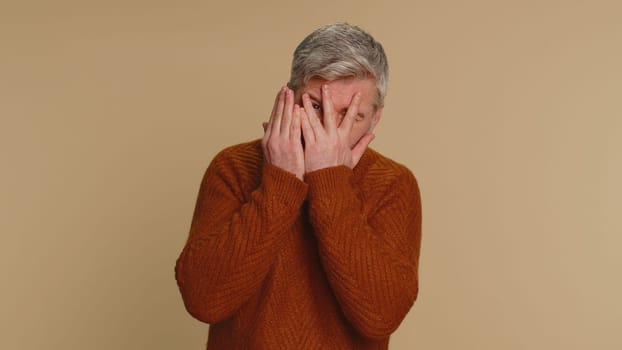 Nosy curious middle-aged man closing eyes with hand and spying through fingers, hiding and peeping binocular gesture, exploring way seeking something in distance. Senior mature guy on beige background